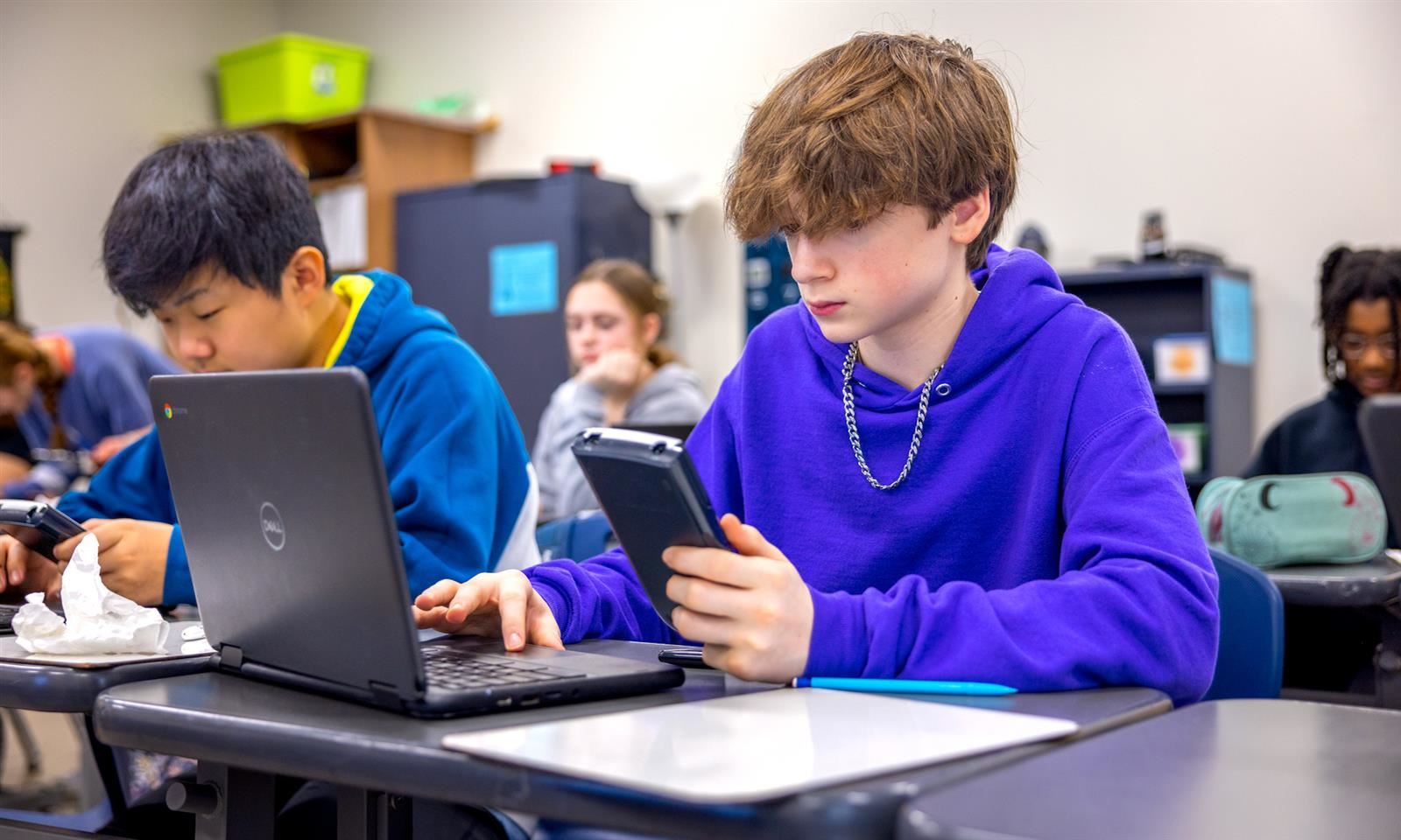 A boy sits at a laptop while holding a calculator